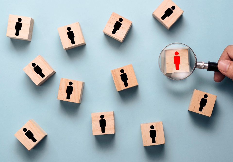 [Pra todos verem] Imagem mostra cubos de madeira com silhuetas de pessoas sobre superfície azul. Mão segura lupa sobre o único cubo com silhueta vermelha