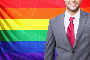 Pra cego ver: Imagem traz bandeira do movimento LGBTQIA+ ao fundo e executivo em meio corpo, à frente, mostrando parcialmente o rosto.