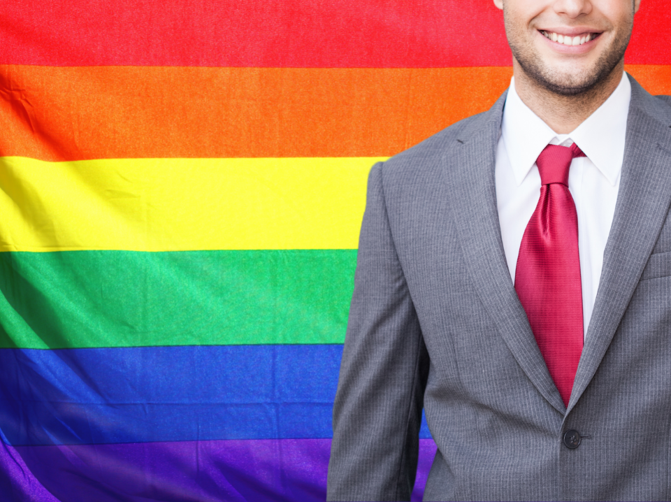 Pra cego ver: Imagem traz bandeira do movimento LGBTQIA+ ao fundo e executivo em meio corpo, à frente, mostrando parcialmente o rosto.