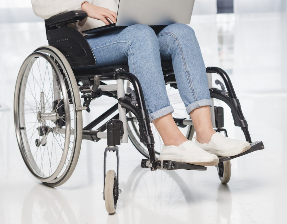 [Pra cego ver] Imagem traz recorte de foto mostrando pessoa com deficiência em uma cadeira de rodas, usando um notebook apoiado sobre as pernas. Destaque está no colo e membros inferiores da pessoa, além das rodas da cadeira.